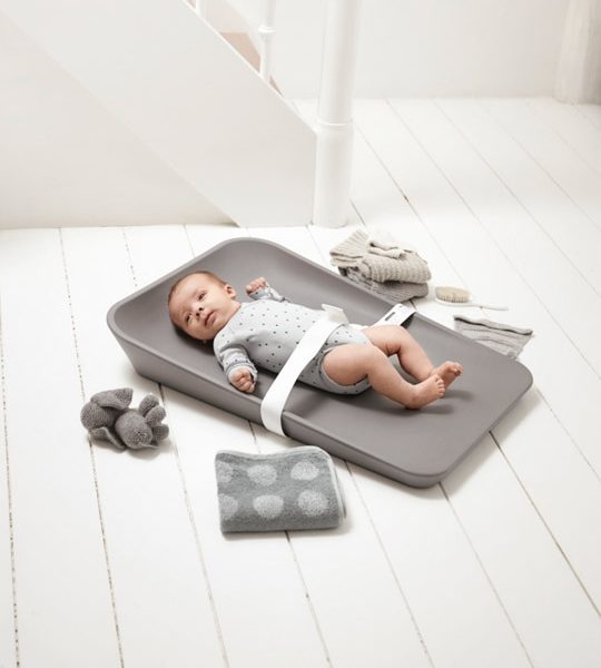Matty Changer in grey with a baby strapped in with a white strap on a white wooden floor with grey folded towels